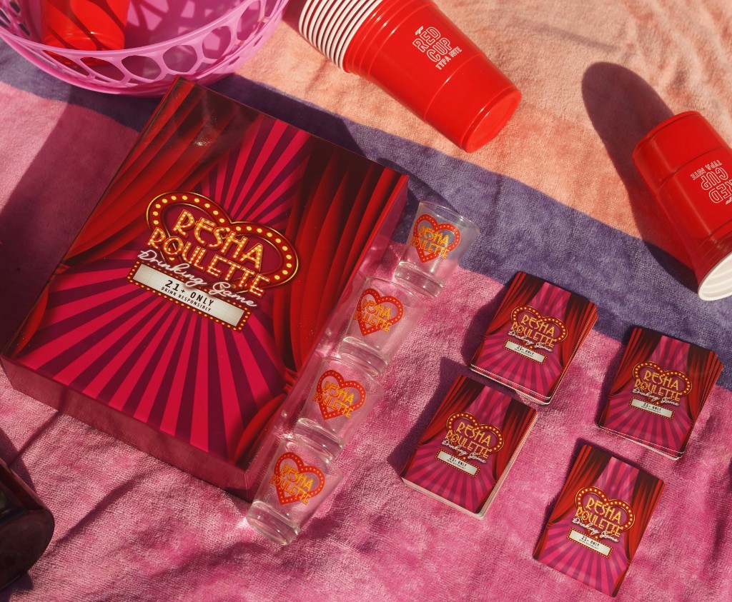 Resha Roulette drinking game: box, shot glasses, and card decks on a striped towel with red cups in bright sunlight.