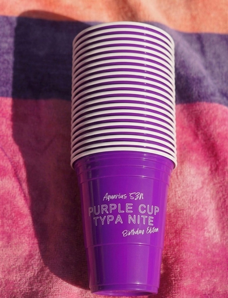 Purple Cup Typa NITE Aquarius Birthday Edition on a pink, orange, and purple striped towel in bright sunlight.