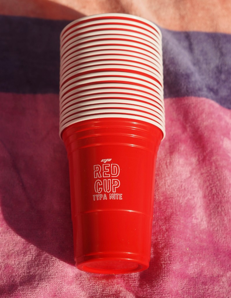 Stack of branded Red Cup Typa NITE plastic party cups on a pink and purple striped towel in sunlight.