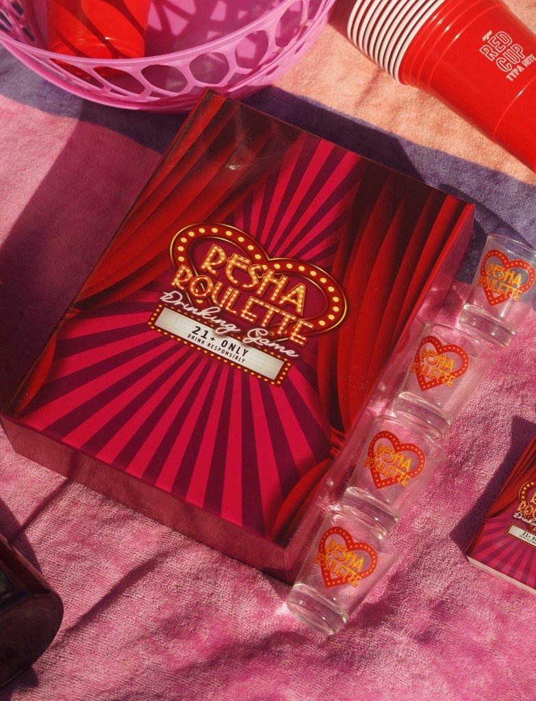 Resha Roulette: game box, branded shot glasses, and red party cups on a pink striped towel.